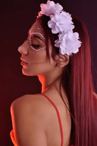 Side profile of a woman with red hair and white floral headpiece wearing pearl face gems in moody lighting.