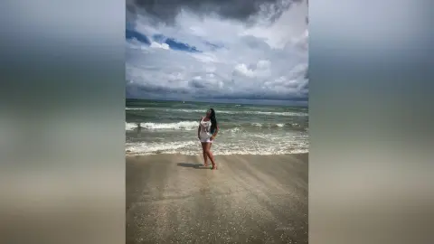 MissyJolie stands on a sandy beach wearing a white bodysuit against a cloudy ocean backdrop.