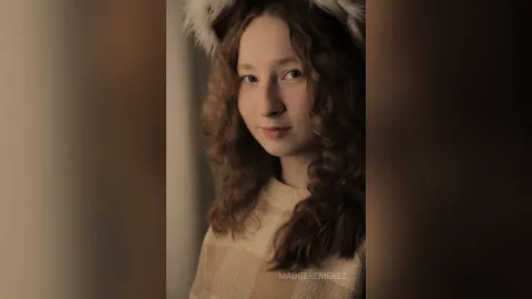 Young woman with curly hair wearing a cream sweater in a warm, moody indoor setting.