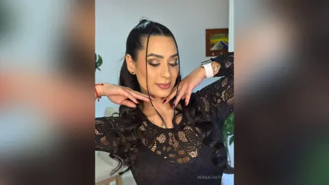 Glamorous woman in a black lace outfit with braided hair and elegant makeup posing in a bright room.