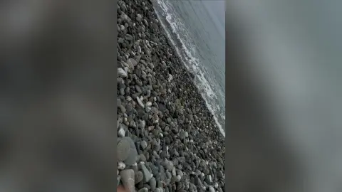 Aerial view of smooth grey pebbles on a rocky shoreline meeting the calm blue ocean waves under soft daylight.