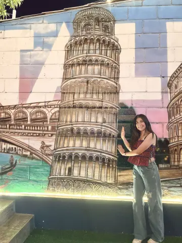 Zharid_Parker smiles while posing next to a large mural of the Leaning Tower of Pisa.