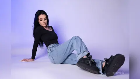 Young woman in black long-sleeve shirt and light blue jeans sitting on a studio floor with a moody purple backlight.