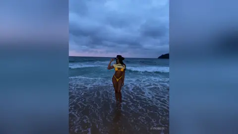 Woman in yellow bikini and black hat standing in ocean waves under a moody twilight sky.