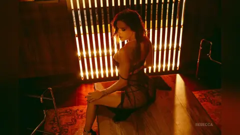 Rebecca sits on a wooden platform in a sheer black lace bodysuit, looking over her shoulder in warm, moody lighting.