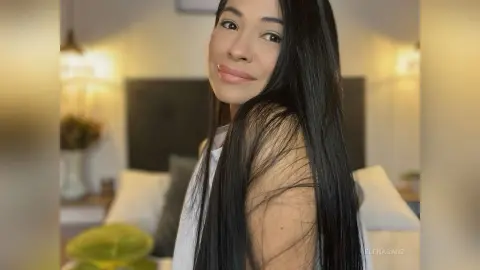 Smiling woman with long black hair wearing a white tank top in a cozy, warm-lit living room.