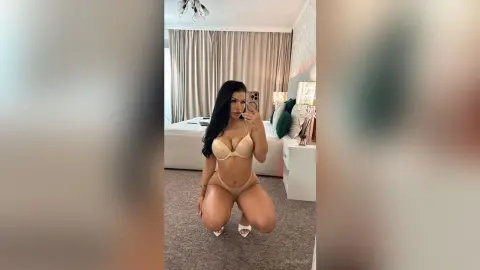 AlmaKane — Brunette woman in beige lingerie posing in a bright, modern bedroom for a mirror selfie.