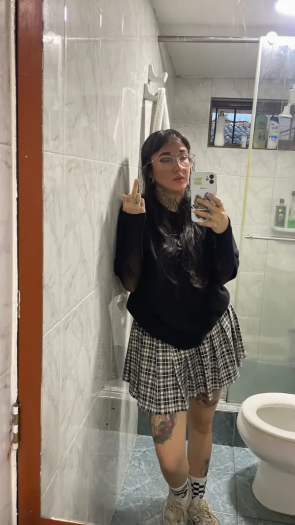 SallyLynn18 — SallyLynn18 takes a mirror selfie in a bathroom, wearing a black long-sleeve shirt and a plaid skirt while showing her middle finger.