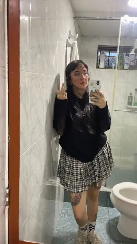 SallyLynn18 takes a mirror selfie in a bathroom, wearing a black long-sleeve shirt and a plaid skirt while showing her middle finger.