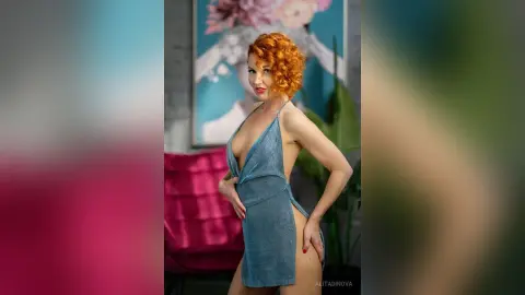 Redheaded woman in a glamorous blue cutout dress posing confidently in a modern living room.
