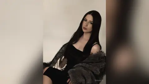 Elegant woman in a black dress and grey faux fur coat posing against a neutral studio background.