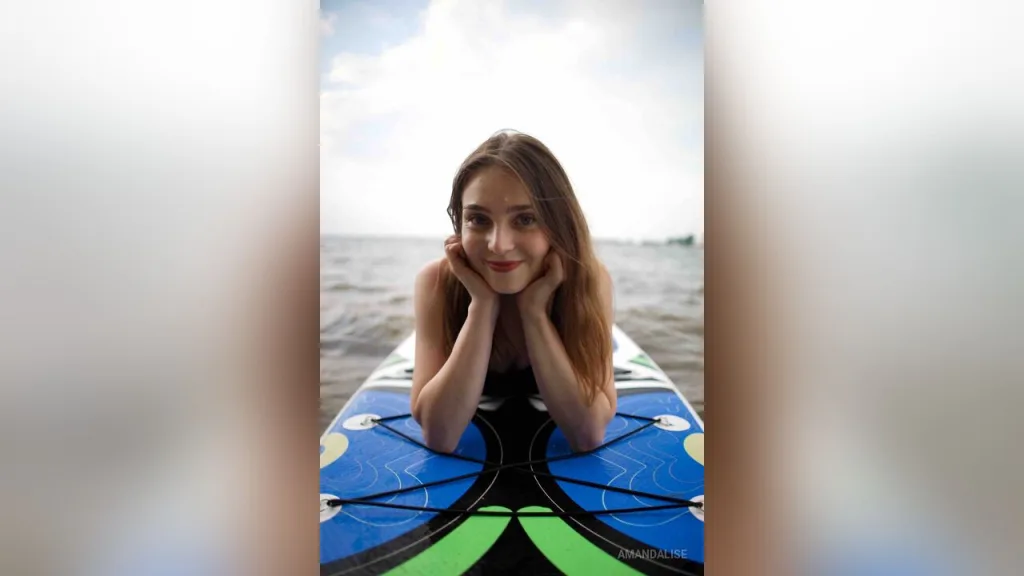 AmandaLise — AmandaLise lies on a paddleboard in the ocean, resting her chin on her hands and smiling at the camera.