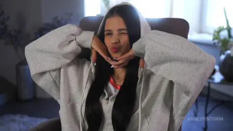 Playful young woman in a grey oversized hoodie winking and making a kissy face while sitting indoors.