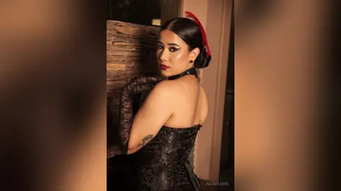 JuliahLewis poses with a bare shoulder and back, wearing a black lace corset and a black choker against a textured wall.