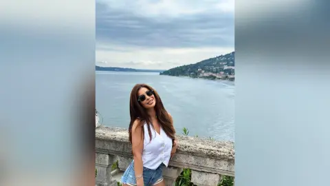 ReynaGomes poses outdoors in a white sleeveless top and denim shorts, wearing sunglasses with a lake and hills in the background.