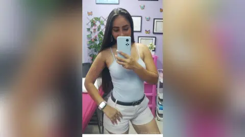 Smiling woman in a white tank top and shorts taking a selfie in a bright, modern bedroom.