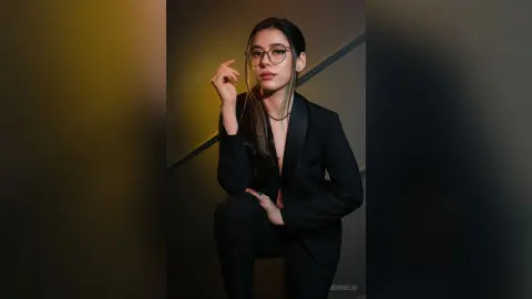 JessieLiu wears a black blazer and matching trousers while posing with one hand near her face and her hand resting on her knee.