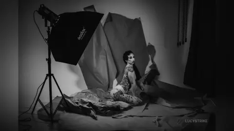 Moody black and white studio portrait of a tattooed woman sitting on crumpled paper backdrop near a softbox light.