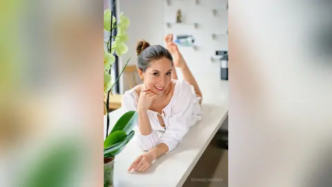 Smiling woman in a white blouse relaxing on a white counter next to a green orchid in a bright modern room.