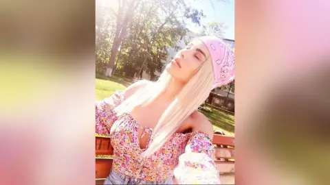 Blonde woman in a floral puff-sleeve top and pink headscarf relaxing outdoors in sunny garden setting.