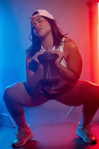 Curvy woman in gym wear performing squats with a dumbbell under dramatic blue and red studio lighting.