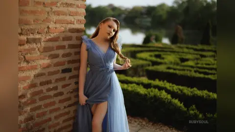 RubySoul poses against a brick wall in a long, blue tulle dress with a high slit, standing in a garden setting.
