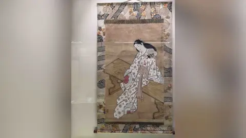 MnemoZeina sits on a wooden bench wearing a white patterned kimono with a red floral detail, looking down with long dark hair.