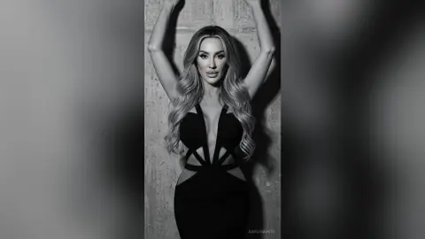 Glamorous blonde woman in a black cutout evening gown posing against a textured wall in a moody black and white portrait.