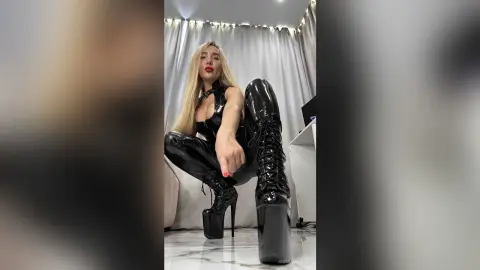 Blonde woman in sexy black latex bodysuit and platform boots posing in a bright modern room.