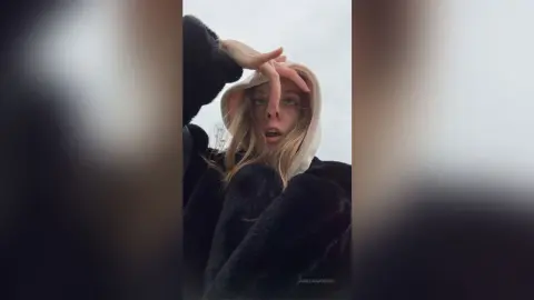 Young woman in a cozy white hoodie and black faux fur coat posing playfully outdoors on a cloudy day.