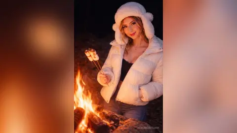 Blonde woman in a white puffer jacket and fuzzy hat roasting marshmallows over a cozy nighttime campfire.