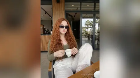 Redheaded woman in a green top and white pants enjoying coffee at an outdoor cafe patio.