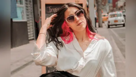 Stylish woman in white button-down shirt and sunglasses posing on a sunny city street with pink hair highlights.