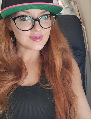 Anna_OG wearing a black tank top and a pink and green cap, showing off her long red hair and glasses while sitting in a chair.