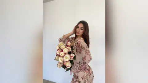 Elegant woman in a floral pink dress holding a large bouquet of white and pink roses against a white wall.