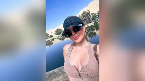Smiling woman in a beige ribbed tank top and black visor enjoying a sunny day by a scenic lake.