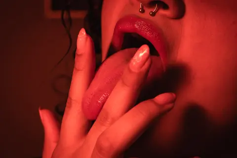 Close-up of a woman with a nose ring sticking her tongue out in moody red lighting.
