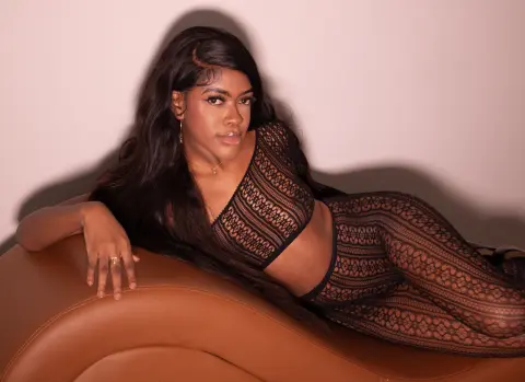 Beautiful Black woman in a black lace two-piece outfit lounging on a brown leather sofa in soft lighting.