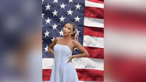 Katia poses in a white sundress with her hands on her hips in front of an American flag.