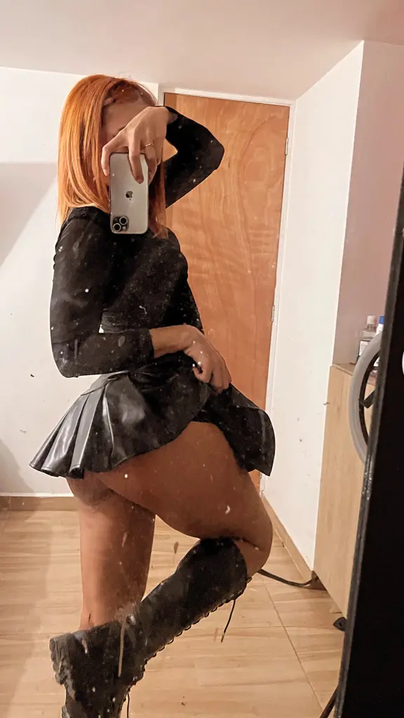 RyleeWilson poses outdoors at night, wearing a black crop top and black leather shorts that show off her legs and red hair.