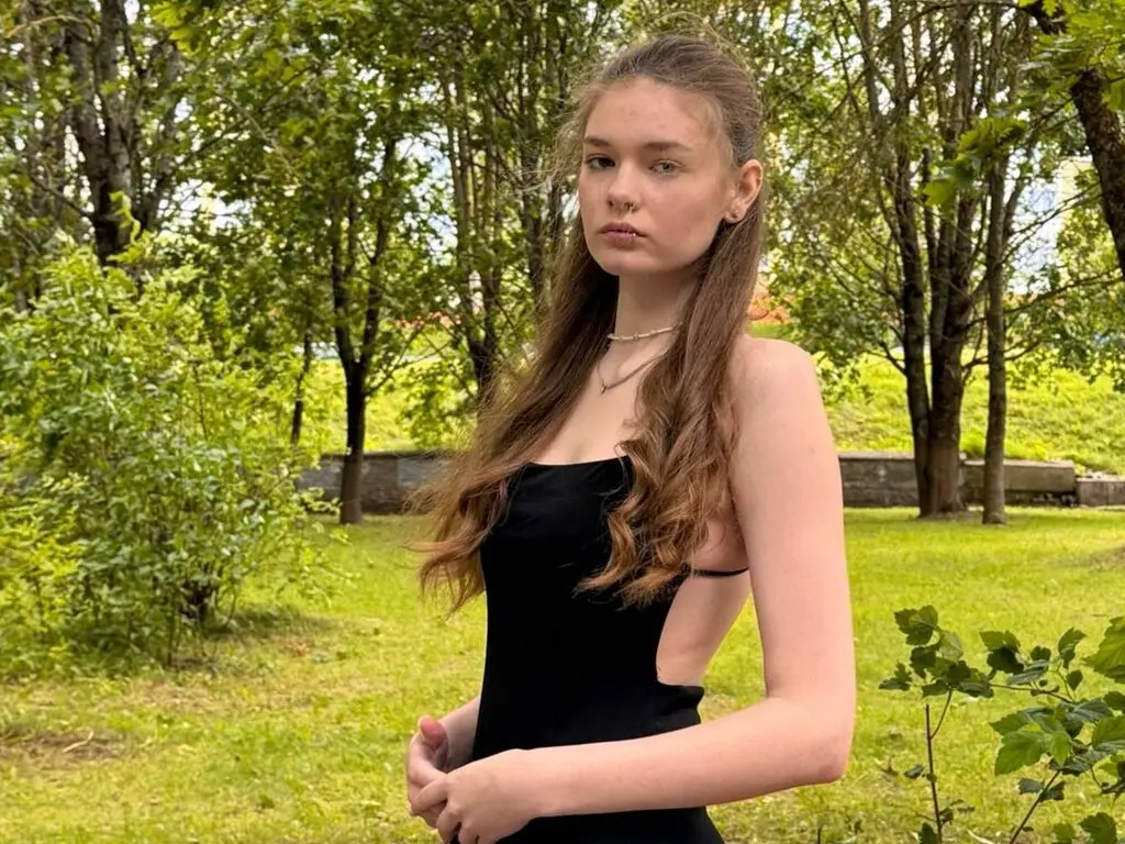 PerryDiotte poses outdoors in a black tank top, showing her long wavy brown hair and neck against a background of green trees and grass.