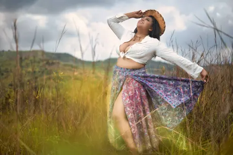 Joyful woman in white blouse and floral maxi skirt posing in a sunny grassy field under a cloudy sky.
