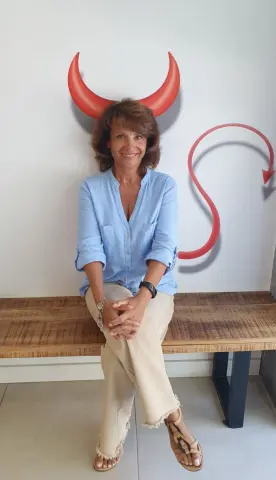 Smiling woman in a light blue linen shirt and beige trousers sitting before a red devil horn wall decal.