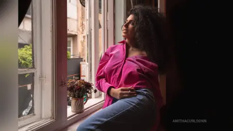 Young woman with curly hair in a pink shirt sitting by a window in soft natural light.