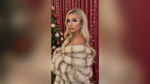 Glamorous woman in a luxurious faux fur coat posing by a decorated Christmas tree in a festive holiday setting.