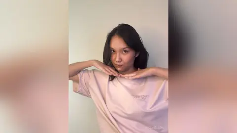 ViolaEbbing poses with her hands near her face, wearing a loose pink t-shirt against a plain white background.