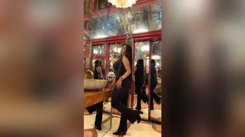 Elegant woman in a chic black sleeveless outfit posing in a luxurious mirrored room with warm lighting.