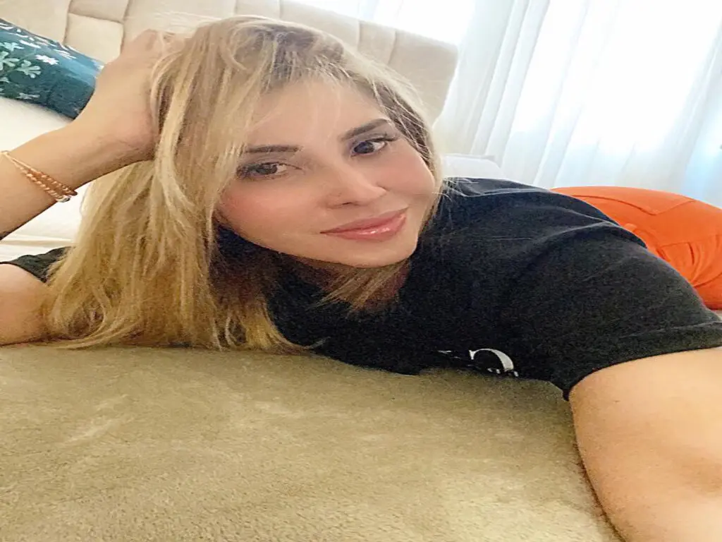 Smiling blonde woman in a black t-shirt relaxing on a bed in a bright, cozy bedroom setting.