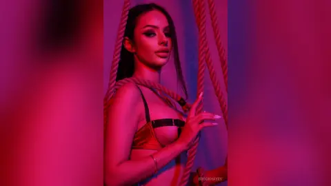 Glamorous woman in gold lingerie posing amidst red ropes under moody pink studio lighting.