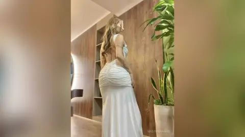 Elegant woman in a white backless evening gown posing in a modern minimalist room with indoor plants.
